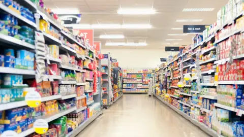 Getty Images Shelves in a grocery store aisle.
The products are blurred out but colourful and the titles at the top of the aisle are blue and "fish and meat" sign is visible.
The floor is cream and the lights are bright.