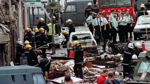 PA Media Firefighters and police officers attending to the aftermath of the Omagh Bomb. There is a white car amongst the wooden and metal rubble, with a fire engine, another black car and van in the background.