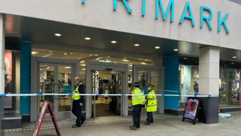 BBC Two police officers and a security guard stand behind police tape at the entrance to a Primark shop