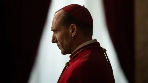Focus Features Ralph Fiennes standing side on in his role as a cardinal in the film Conclave. He is wearing red robes and a skull cap. Behind him is a window with parted curtains. He is closing his eyes in prayer.