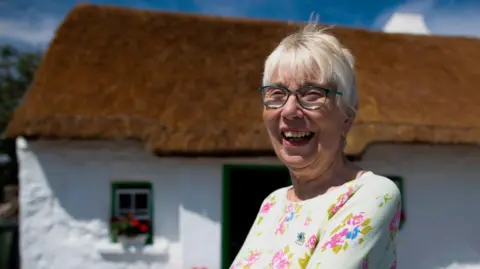 PAUL FAITH/AFP via Getty Images Margaret Gallagher pictured smiling outside her thatched cottage in July 2019. She has short grey hair and glasses and is wearing a yellow floral top.