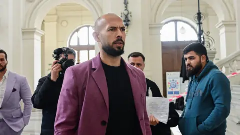 Getty Images Andrew Tate wears a purple jacket as he walks inside a court in Bucharest, as a photographer behind him takes his picture