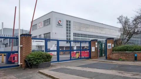 Getty Images The entrance to the assembly plant of the automotive manufacturing company IBC Vehicles Ltd, operating under Vauxhall Luton and producing light commercial vehicles, in December 2021. It shows blue metal gates, red barriers, and a grey building with lots of windows