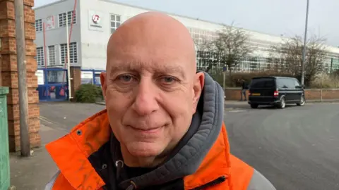 Sam Read/BBC Dave wears a bright orange and grey work anorak and a grey hoodie. He is bald and stands next to the road outside the Vauxhall factory. The Vauxhall emblem is on one corner, and the building recedes into the distance with many windows across its two stories.