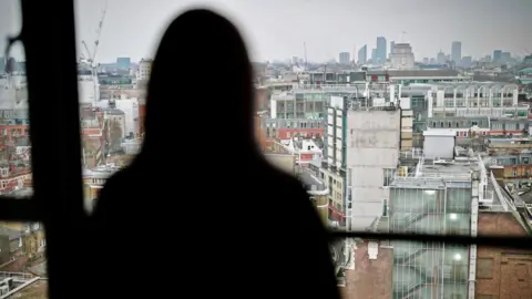 Beth, pictured in a blurred silhouette against a high window, looking out onto tall buildings stretching into the distance on an overcast day