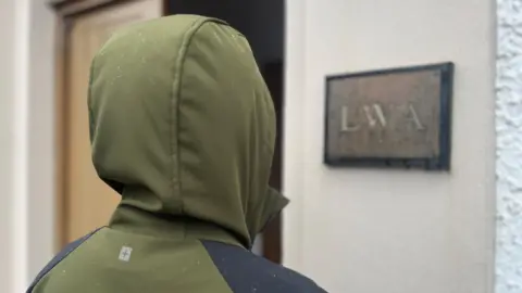 BBC Unidentifiable person with their back to the camera wearing a khaki waterproof jacket with the hood up. They are standing outside a building with a sign that says "LWA".