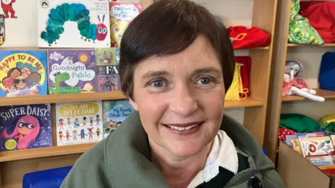 BBC Tracey Roleston smiling at the camera. She has short brown hair and is wearing a green top. Behind her are bookshelves with books and toys.