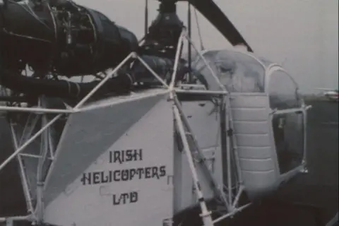 BBC archive A small white framed helicopter is seen from behind with the words 'Irish Helicopters LTD' printed in black lettering on a rear panel. A black engine sits above the frame with the rotor blades slightly visible. To the front is a small cabin which appears like it would have room for only two or three people. It's made from thin metal strips surrounded by large see through panels.  