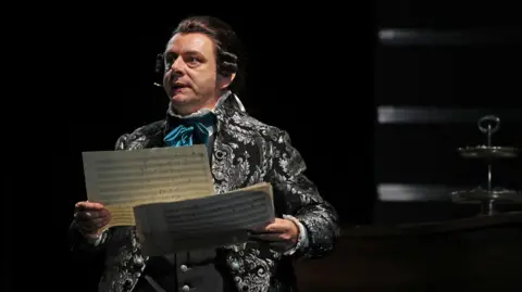 Getty Images A picture of Michael Sheen playing the role of Antonio Salieri during a media call for Amadeus at Sydney Opera House on December 30, 2022 in Sydney, Australia. He is wearing a patterned sliver and black jacket and is holding papers of music. The spotlight is on him.