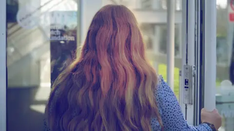 A woman walks through a door of a glass building. You can see the back of her head, she is wearing a blue top with black dots and has long wavy blonde/red hair