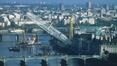 Marks Barfield An elevated view of London and the River Thames with a huge yellow crane lifting the Millennium Wheel into a vertical position