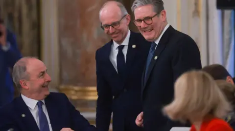 Reuters Left to right:
Micheál Martin is smiling from a seated position at Keir Starmer who is standing and smiling at a woman wearing red who is seated to his left with blond hair to her shoulders with her face turned towards him and away from the camera. Behind Keir, standing is Luc Frieden who is also smiling. Behind the four of them, is a regal looking gold frame with an autumnal coloured pastel painting.
Micheál Martin is wearing a navy suit, white shirt and blue tie. He has short dark grey hair.
Luc Frieden is wearing a navy suit, white shirt and navy tie. He has a bald head with short hair on either side and is wearing black square glasses.
Keir Starmer is wearing a navy suit, white shirt and teal blue tie. He has grey hair brushed to the right and black glasses.
All three of the men are wearing a yellow badge on their suit jackets.
