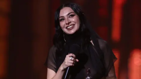 Getty Images Charlie XCX at the podium - again - to receive another Brit. She is wearing a sheer black dress and has her dark hair down.