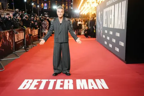 PA Media Robbie Williams wears a dark striped, slightly baggy suit on the red carpet for his film Better Man. Photographers can be seen behind the barrier in the background. Robbie gestures towards the red carpet in front of him which has the words "Better Man" written on it in white letters.