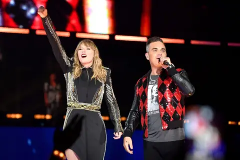 Getty Images Taylor Swift and Robbie Williams on stage together in London in 2018. She wears a black dress with metallic details and he wears a Taylor Swift Reputation top with a red and black sparkly jacket on top.