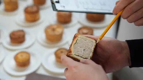 PA Media A photo of someone holding half a pork pie, and a pencil is pointing at it. In the background, underneath are more pies on plates on a table.