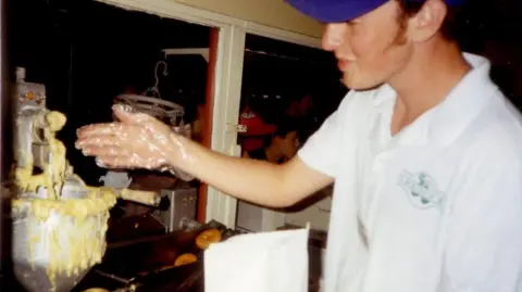 Richard Drummond Richard working at food counter. He is wearing a blue cap and white polo shirt, with a green Oakwood logo on it. He is holding his arm out, and wearing a plastic glove. In the left hand side an electric mixing bowl can be seen with donut batter spilling over the side. Ring donuts can be seen in the background in another piece of equipment.