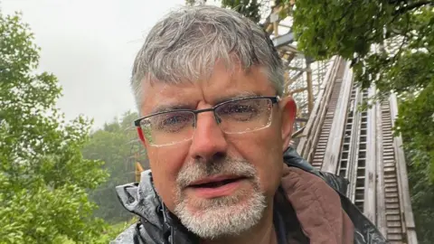 Korey Kieptfer Korey Kieptfer standing on the Megafobia ride on a wet day, he has short, grey hair and a goatee. There are raindrops on the lenses of his glasses and the track can be seen behind him. Trees can be seen on either side of Korey.