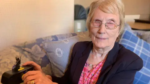A woman with glasses is sitting on a sofa and turning the knob of a radio. She is wearing a navy blazer and a pink colourful top underneath.
