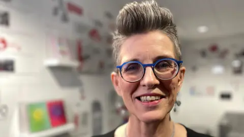 Head shot of Debbie Lockwood who has short, grey spiky hair and is wearing round glasses with a blue frame.