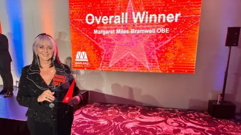 Slimming World Mrs Miles-Bramwell holding a trophy, and behind her is a screen saying "overall winner Margaret Miles-Bramwell".