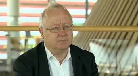 Leighton Andrews looking past the camera wearing a black suit and a white shirt which is open at the collar. He has short white hair and glasses. Behind him the Senedd's distinctive wooden funnel.