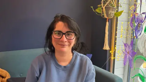 A female customer wearing glasses sat down smiling on a light blue sofa. She has dark hair framing her face and is wearing a blue sweater. On the right hand side a window is decorated with purple and white flowers and fairy lights.