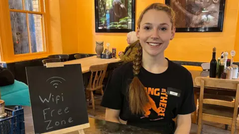 Lili smiling in a cafe by a small chalk board which reads 'wifi free zone'. She has long brown hair, tied into a plait, and is wearing a black t-shirt. Wooden tables can be seen in the background against a bright yellow wall. A customer is sitting at one of the tables.