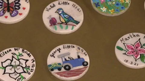 6 white badges with different designs sit on a brown table cloth. The badge on the bottom right is of a pink plant and the date 15/03/21. The badge in the middle has a blue tractor with the name and date, George Little, 16/03/20. Above that is one of a Blue tit and a pink ball of wool with knitting needles, it says Violet Little, 17/01/21. Bottom left is of white flowers and the name Eileen McBride, 19/12/21. Top left has a drawing of a purple and orange butterfly. Top right is of a garden filled with a variety of flowers.
