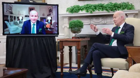 Getty Images Micheál Martin pictured on a TV screen in the White House during a video call with former US President Joe Biden in March 2022. Both men are wearing navy suits, white shirts and green ties. A bowl of shamrock is on a table in front of Biden.