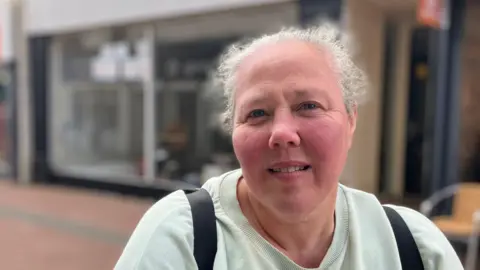 A woman with tied-back grey hair wearing a light green sweatshirt and black backpack and smiling at the camera, with the front of shops behind hr.