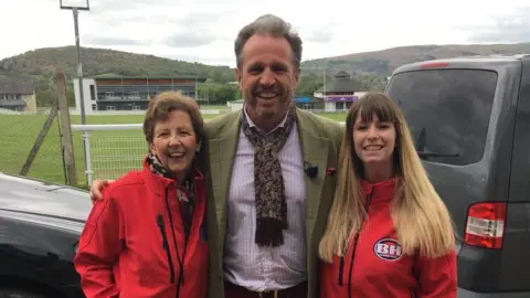 Jane Cooke Jane Cook on the left with short light brown hair in her mid-60s wearing a red Bargain Hunt branded fleece jacket. in the middle is expert Richard Madley wearing a brown and gold Paisley scarf, green tweed-type jacket and small check shirt. Next to Richard is Megan with long blonde hair and also wearing a red Bargain Hunt branded fleece. All three are stood close together between a car and a van with the main ring at the Royal Welsh Showground in Builth Wells in the background. 