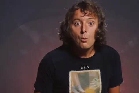 Michael Putland/Getty Images Jasper Carrott, with curly hair, wearing a largely black T-shirt, with ELO named on it.