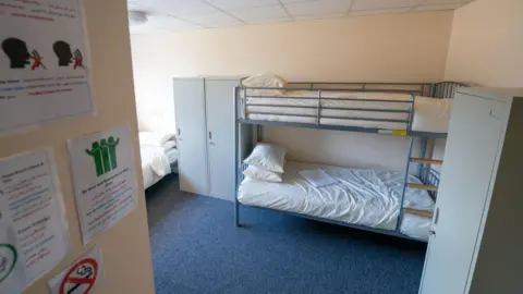PA Media On the right of the image there are bunk beds, that have a metal frame and white pillow and duvet covers. On the left there is another single bed, and a wooden wall with signs on.