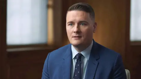 Wes Streeting, who has brown hair and blue eyes, wearing an air-force-blue suit with a navy patterned tie and light blue shirt, sitting in a dark wood panelled room