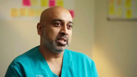 Dr Krishna Kasaraneni, who has a shaved head and a closely trimmed greying beard, wearing turquoise scrubs with Steel City General Practice written on the right chest. He is photographed from the head and shoulders with the background of his GP surgery behind him out of focus.