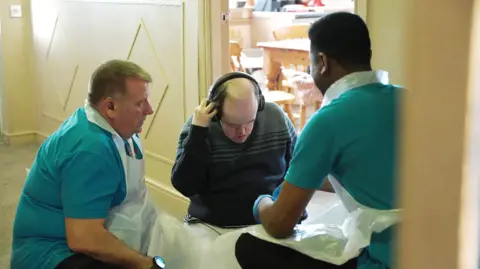 Darren Leigh, who has light brown hair, wearing a teal polo shirt and a plastic apron, kneels down beside his social care client Martin. Martin is looking downwards and holding large headphones against his ears. Darren's colleague is crouching down in front of Martin with his back to camera.