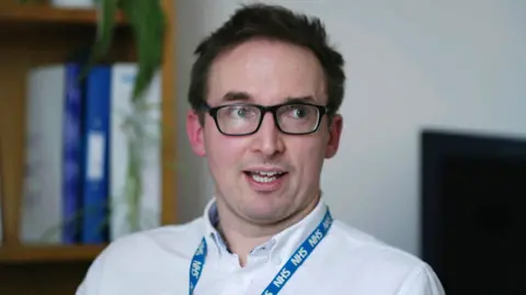 Prof Andy Swift, who has with brown hair and black-framed glasses, wearing a white shirt and an NHS lanyard, pictured in an office with shelves behind him