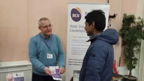 Bedford Credit Union A man in a blue jumper, handing out a leaflet at an event, standing by a banner. The man he is talking to is smiling and has on a blue coat, and is looking to the left. There is a plant to the right.
