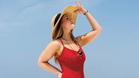Hannah Howie Hannah Howie, who has long, blonde hair and is wearing a red dress with white spots, strikes a pose against a clear blue sky. Her right hand is on her hip and he left hand is tipping the top of her straw hat.