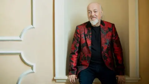 Chatterbox Media Comedian Bill Bailey wears a red floral blazer, black t-shirt and trousers. He sits leaning against a cream coloured wall and smiling.