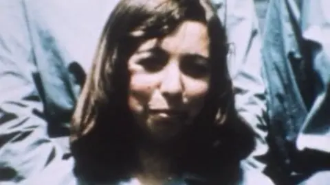 Handout A blurred photograph of Lesley Whittle. She has shoulder-length brown hair and is smiling at the camera. Behind her are people wearing blue tops but we cannot see their hands or faces due to how close they are to Lesley.