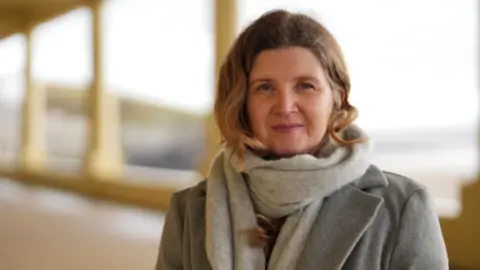 BBC News Johanna Robinson looks towards the camera with a neutral expression. She wears a grey coat and scarf and has her blonde hair tucked behind her ears.