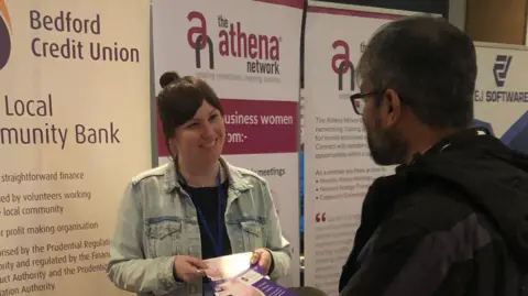 Bedford Credit Union Katrina Allen, holding a leaflet, smiling at a man, in front of a number of banners. She is wearing a dark top, blue denim jacket, with a blue lanyard round her neck. Her long dark hair has been put up. Another man has his back to her, and has glasses and a beard and is wearing dark clothes.