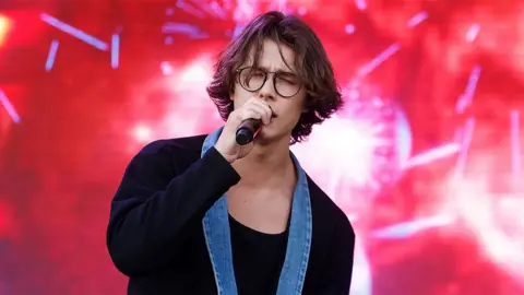 Getty Images David Kushner singing into a microphone with his eyes closed wearing a black jacket with a denim collar