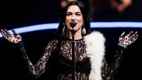 Getty Images Dua Lipa sings into a microphone while on tour. She is wearing a black lace top and large pendant earrings, with a white fake fur stole draped across her left shoulder.