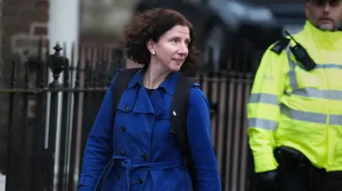 PA Media Anneliese Dodds is seen leaving a cabinet meeting, she is wearing a blue coat and backpack. A security officer in high vis is in the background.