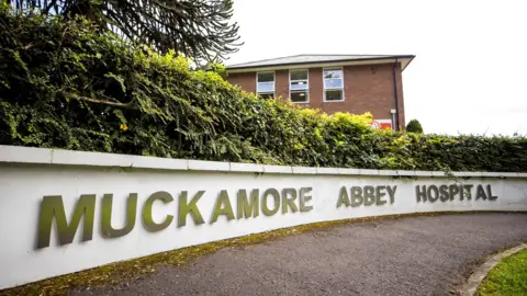 PA Media A white wall reads 'Muckamore Abbey'. There's a red brick building behind the low wall.  A small green bush runs along the top of the white wall. 