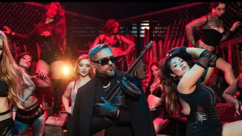 Instagram/Yo Yo Honey Singh A picture of Yo Yo Honey Singh wearing a black jacket over a black suit and a silver chain with black leather gloves. He is holding a gun and has background female crew wearing black dresses