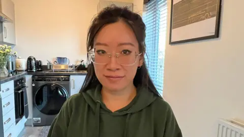 BBC A woman in a kitchen looks at the camera with a neutral expression. She wears frameless glasses and a green hoodie and her dark hair hangs behind her shoulders. Behind her is a washing machine to the left and a vase of yellow flowers is on the worktop beside it.

